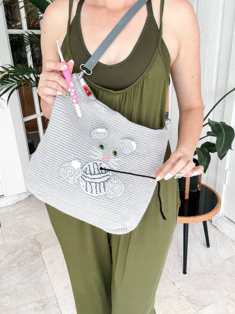 Person wearing a green outfit holding a gray woven bag with a bear and yarn design, perfect for carrying a cat crochet project bag pattern, and a pink toothbrush in the other hand. A plant is visible in the background.