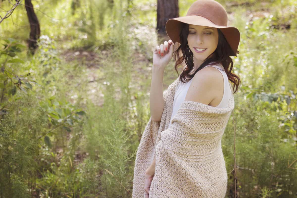 A woman wearing a brown hat and crochet wrap in a field of greenery.