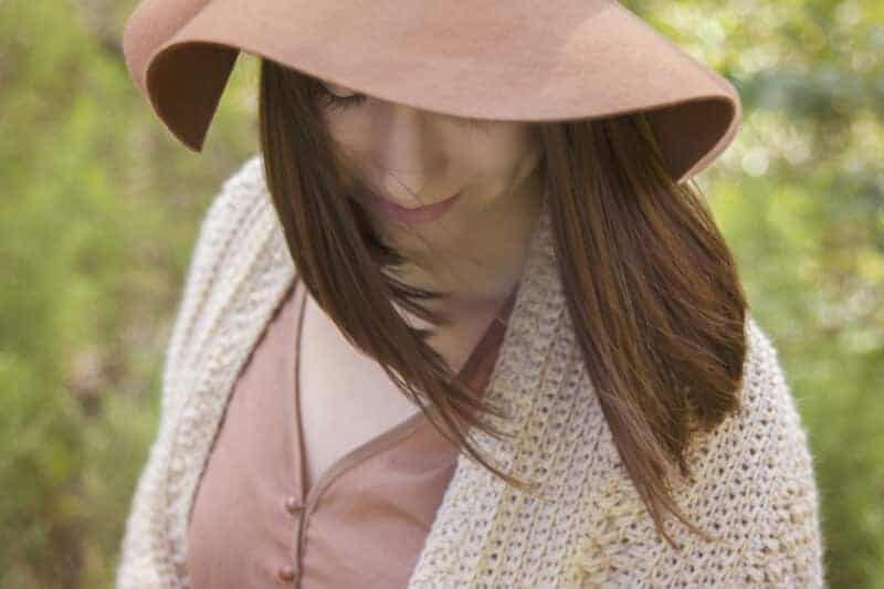 A woman wearing a brown hat and crochet wrap in a field of greenery.
