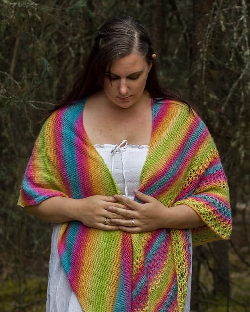 A woman in a colorful knit shawl and white top standing in a wooded area, looking down thoughtfully with her hands clasped.