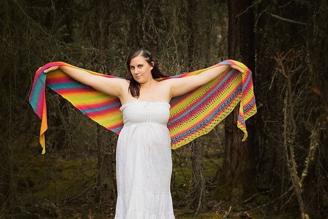 A woman in a white dress standing in a forest, holding a light summer shawl outstretched with her arms.