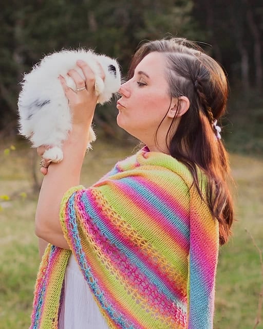 A woman in a colorful light summer shawl kisses a small white fluffy rabbit she is holding outdoors.