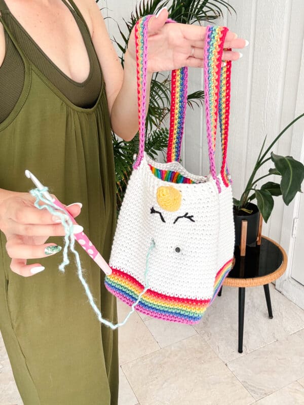 A person in a green dress holds a crochet hook and a partially crocheted unicorn-themed project bag with colorful stripes. A table and plant are in the background.