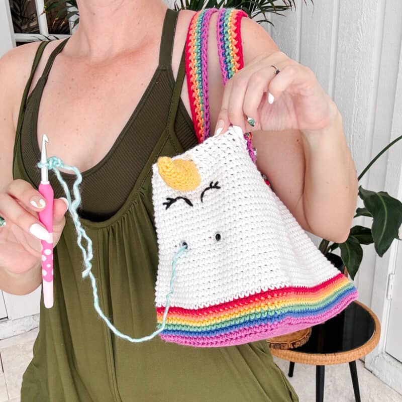 Person holding a white knitted unicorn bag with rainbow stripes and a unicorn design, perfect for storing supplies as a crochet project bag, along with a crochet hook and light blue yarn.