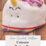A white crocheted unicorn project bag with a yellow horn, black eyes, and rainbow details. Text reads: "Free Crochet Pattern - Unicorn Project Bag - step-by-step - www.brianakdesigns.com. Perfect for your next crochet project bag creation!