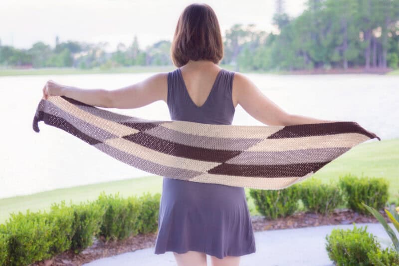 A woman stands facing a lake, holding out a colorwork crochet shawl with each hand, dressed in a sleeveless grey dress.