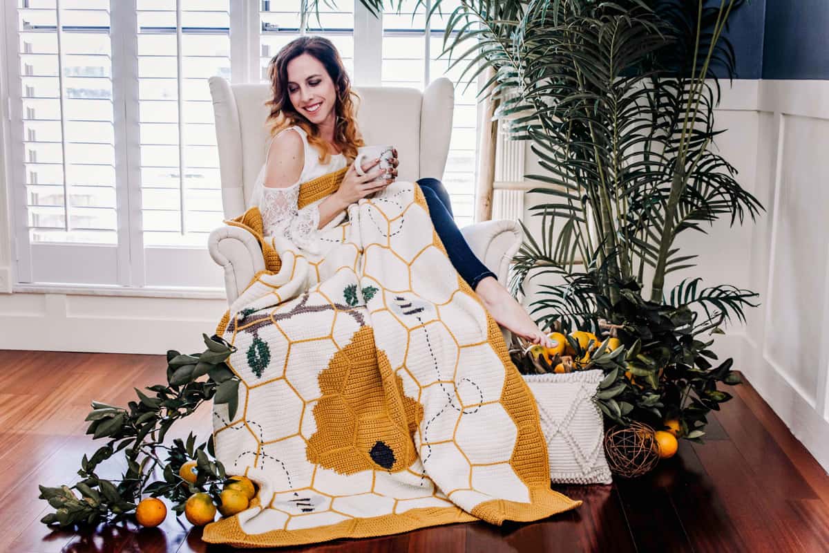 A woman sitting in a large chair wrapped in a bee blanket, holding a mug, with scattered oranges and greenery around her.