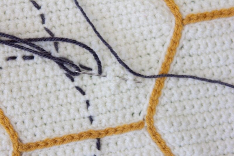 A close-up of a needle with thread on a crocheted Bee Blanket with white squares and yellow borders.