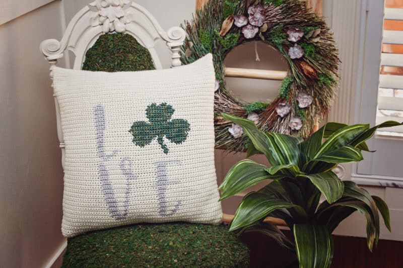A St. Patrick's Day crochet shamrock pillow is sitting on a chair in front of a wreath.