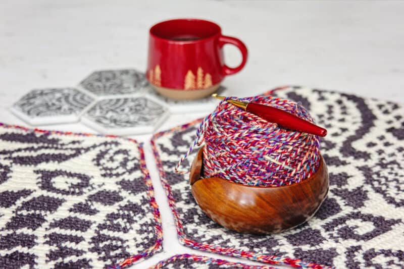 A wooden yarn bowl with knitting needles and multicolored yarn on The Gaudi Sidewalk Blanket, with a red mug and coasters in the background.