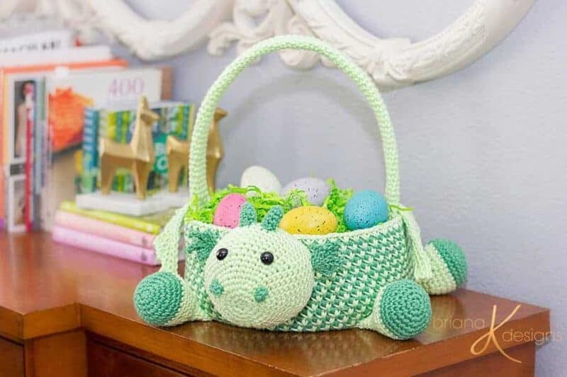 A crocheted dinosaur easter basket sitting on top of a bookshelf.