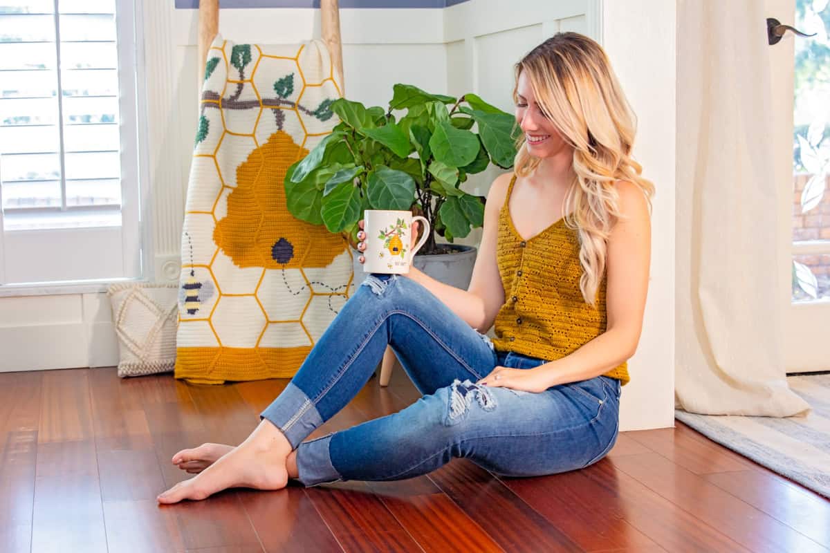 A woman in a yellow top and ripped jeans sitting on a hardwood floor, holding a mug, smiling, with a large plant and a honeycomb crochet hanging in the background.