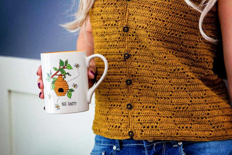 A blonde woman in a mustard honeycomb crochet top holds a white mug with a "bee happy" design featuring a beehive and flowers.