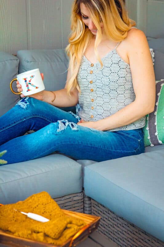 A woman sitting on a couch, wearing a grey sleeveless top and ripped jeans, holding a mug, with a notebook on how to honeycomb crochet laid out on a wooden tray next to