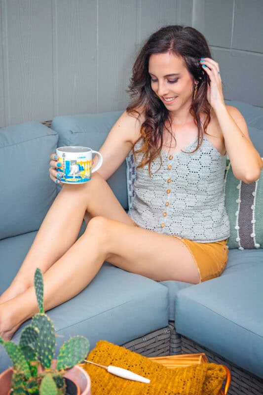A woman sits on a gray sofa holding a mug, smiling, with a potted cactus and a "How to Honeycomb Crochet" book beside her.