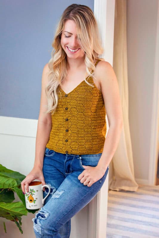 A woman smiling and holding a coffee mug, wearing a mustard honeycomb crochet top and blue jeans, leaning against a wall indoors.