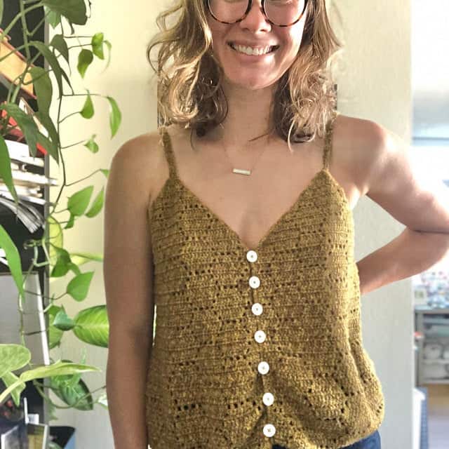 A smiling woman wearing glasses and a handmade mustard-colored honeycomb crochet top with white buttons, standing indoors.