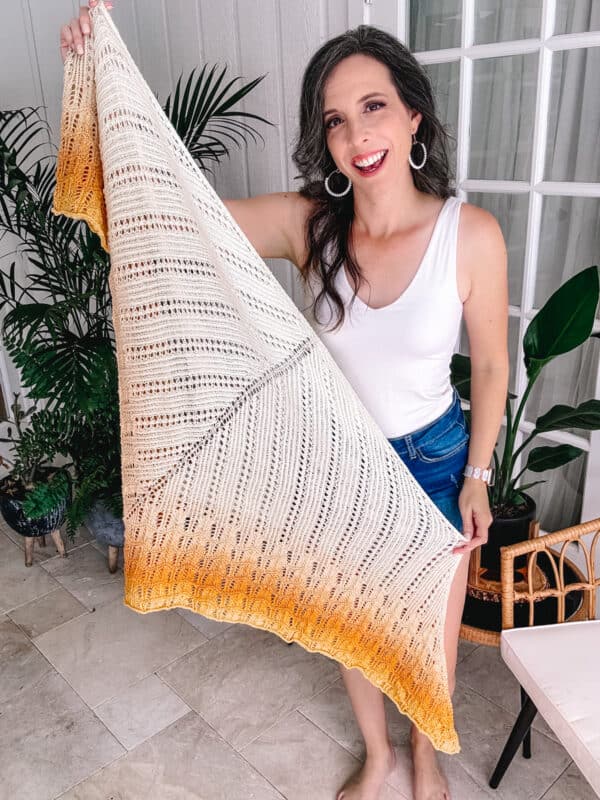 A woman in a white top and jeans holds up a large, hand-crocheted Ray of Sunshine Knit Shawl, smiling in a bright, plant-filled room.