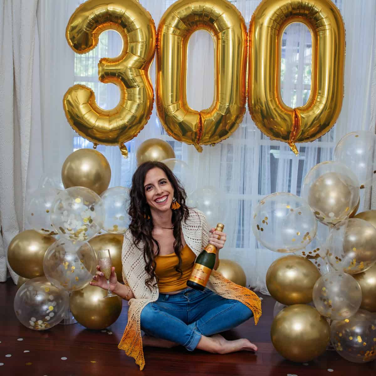 A woman sitting on the floor smiles, holding a champagne bottle, surrounded by gold and white balloons and "300" gold balloons in the background, dressed in a Ray of Sunshine Knit Shawl.