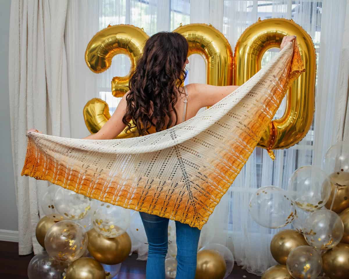 Woman with long curly hair holding up a Ray of Sunshine Knit Shawl, standing in front of gold "2018" balloons and gold and white balloons on the floor.