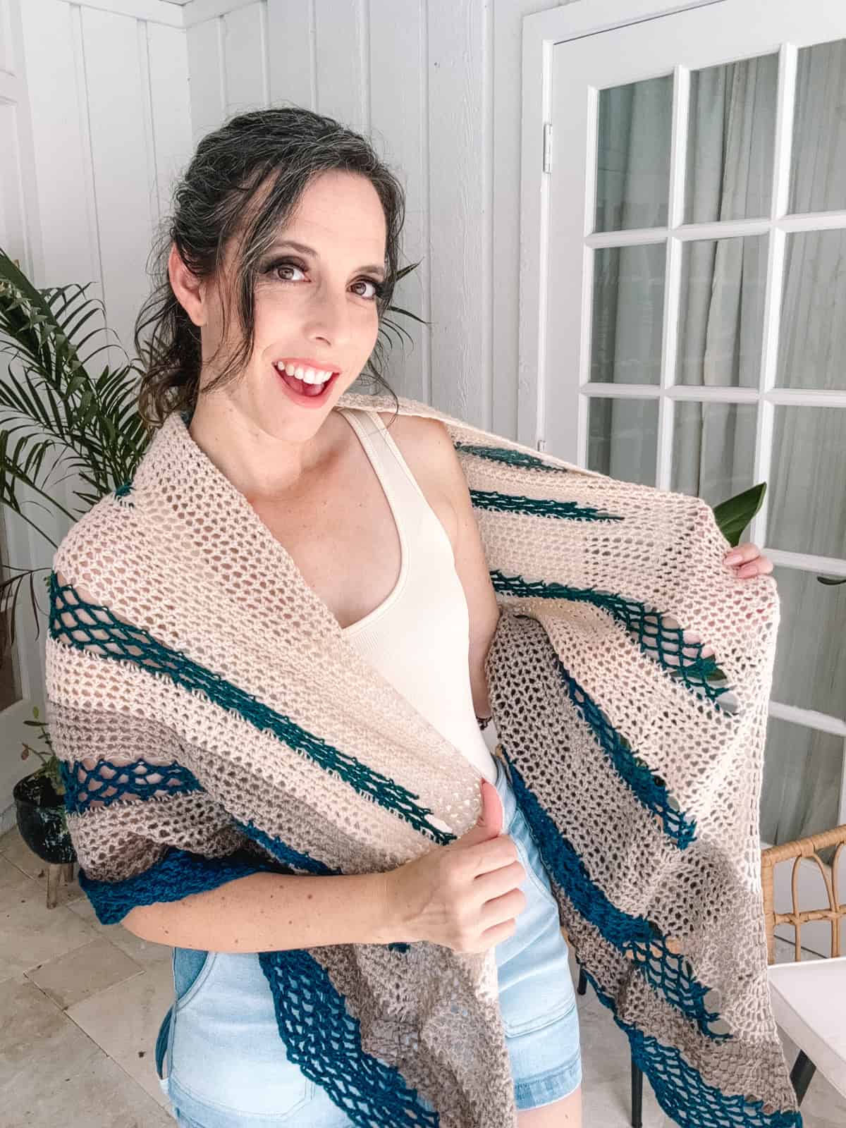 Woman indoors smiling, wearing a light top and shorts, holding an open Fisherman's Wharf Crochet Shawl adorned with blue and beige patterns.