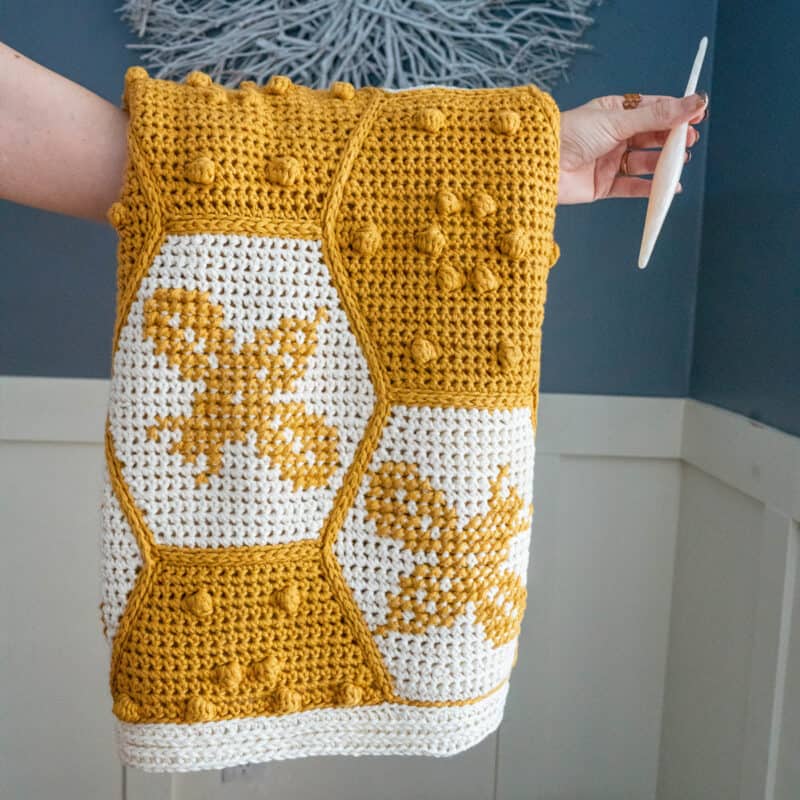 A person holding The Honey Bee Blanket with a bee design and a crochet hook.