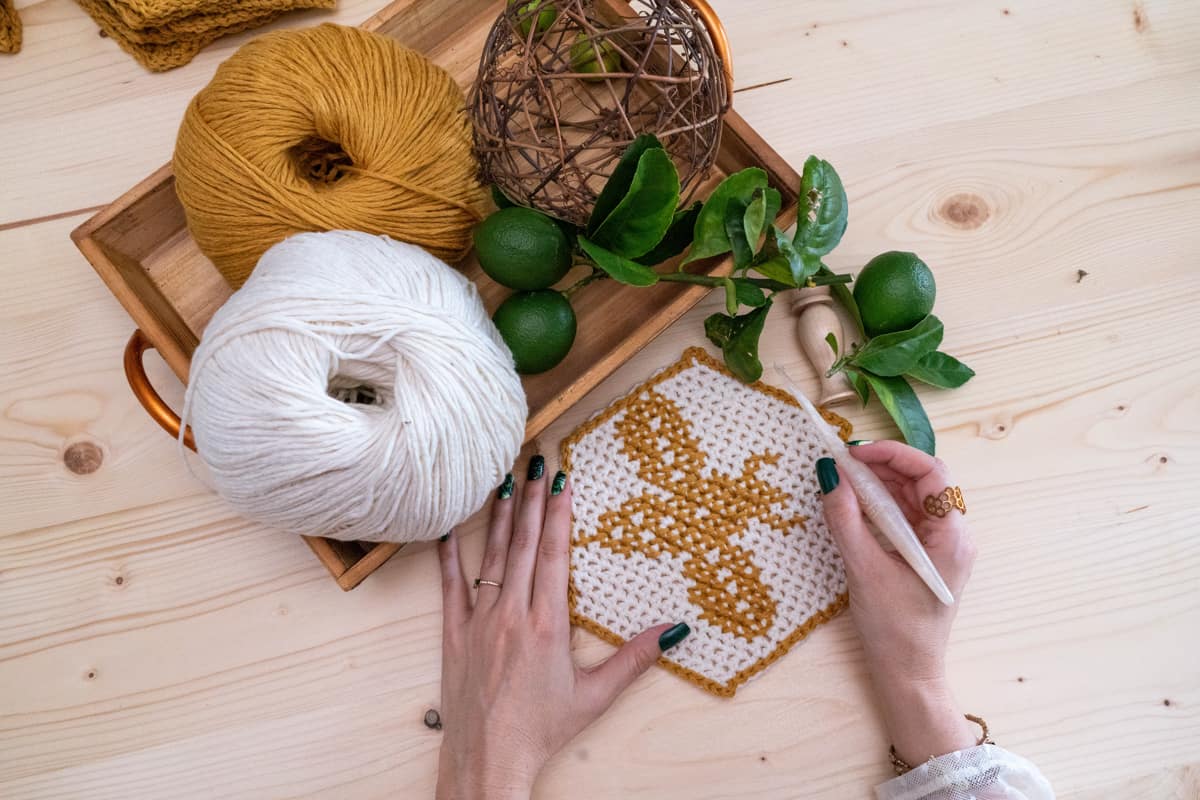 Person crafting The Honey Bee Blanket with yarn and crochet needle on a wooden table with plants and yarn balls nearby.