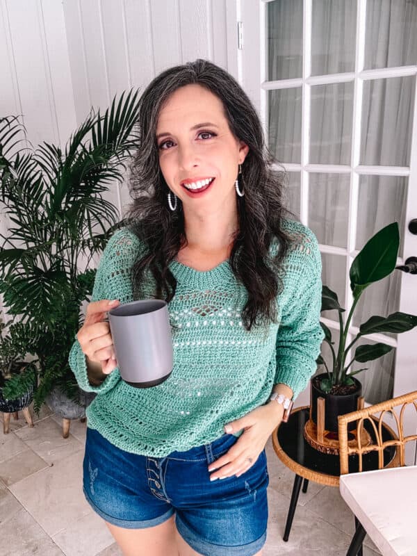 Woman in a sagebrush Lindy chain sweater and denim shorts smiling and holding a gray mug in a bright sunroom.