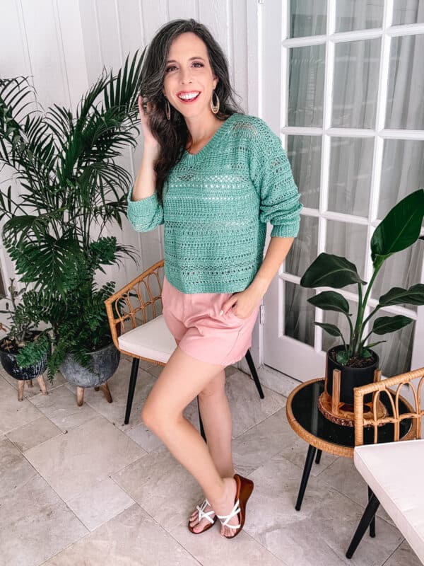 A woman in a sagebrush green crochet top and pink shorts smiling while posing in a sunroom with potted plants and rattan furniture.