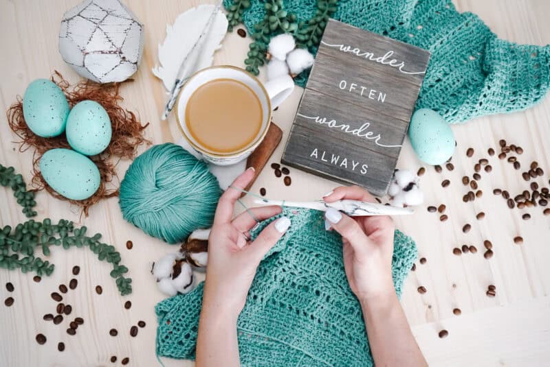 Hands crocheting a Sagebrush Lindy Chain Sweater near a cup of coffee, surrounded by blue speckled eggs, beads, and a wooden sign with the text "wander often wonder