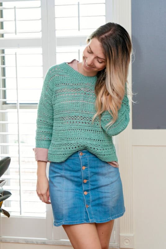 A woman with long highlighted hair, smiling gently, wearing a sagebrush crochet top and a blue denim skirt, standing in a room with shutters.