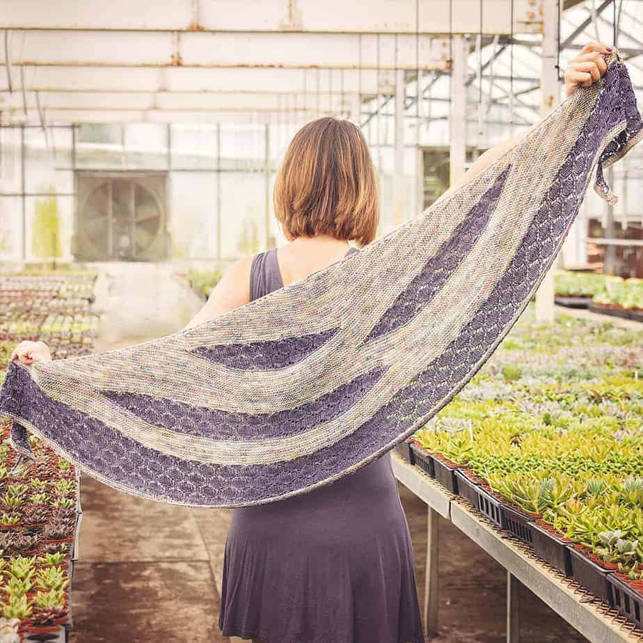 A woman wearing a honeycomb knit shawl in a greenhouse.