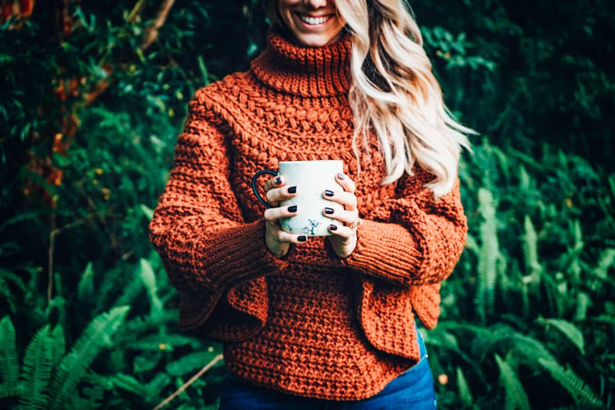 A woman holding a coffee cup in a crocheted poncho.