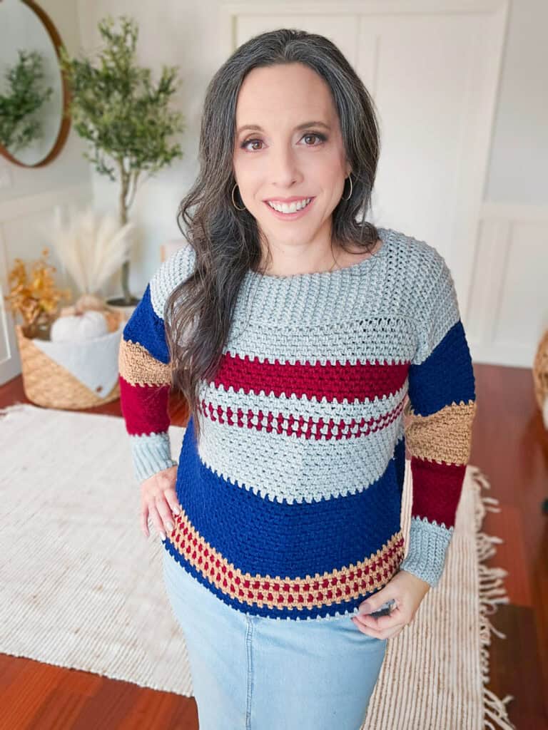 A person is standing indoors, wearing a vibrant Gingersnap Crochet Sweater with red, blue, and beige stripes paired with a light blue skirt. They have long dark hair and are smiling at the camera.