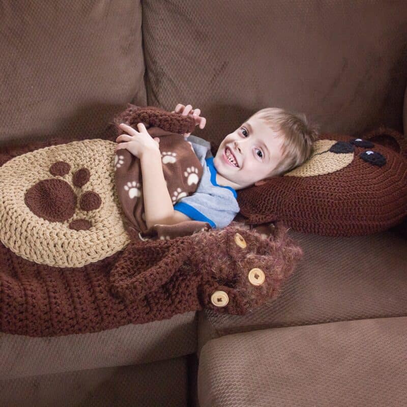 A young boy snuggles on a couch with a crocheted teddy bear in a cozy sleeping bag.