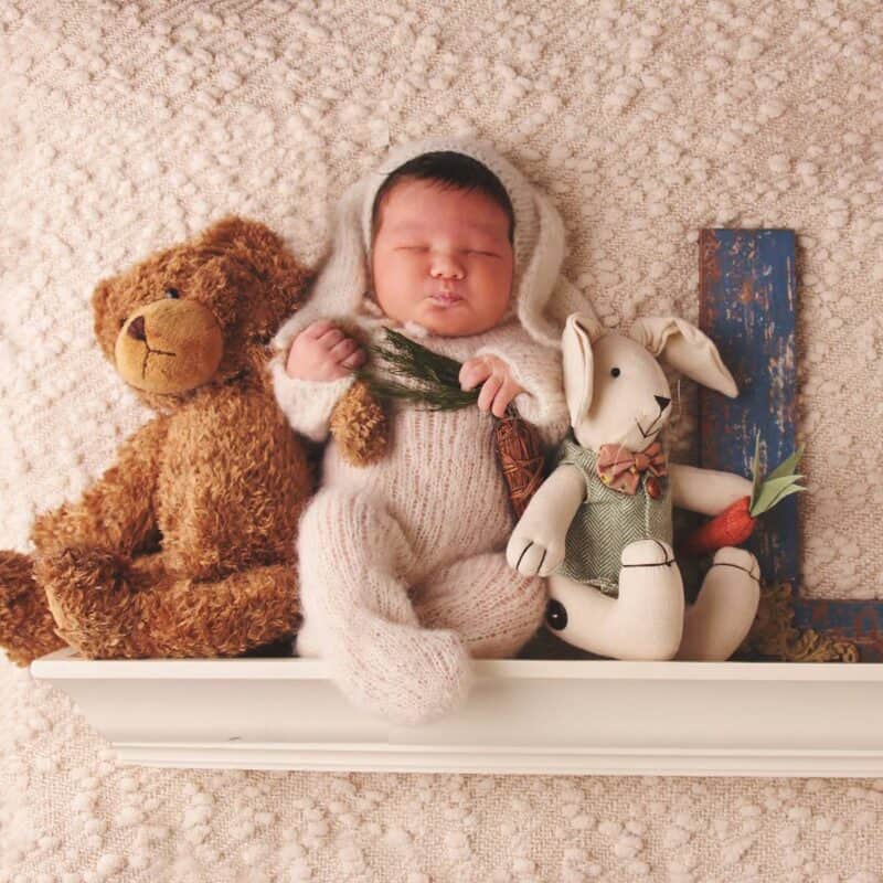 A baby is perched on top of a shelf surrounded by cute teddy bears and a soft knitted bunny.