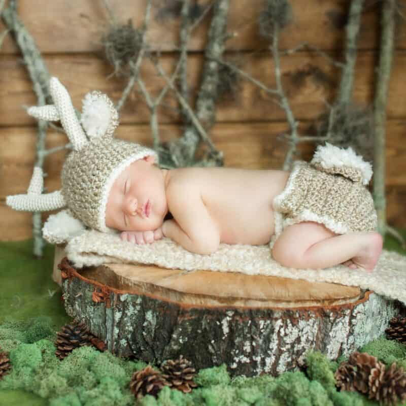 A newborn baby in a crocheted deer outfit resting on a stump.
