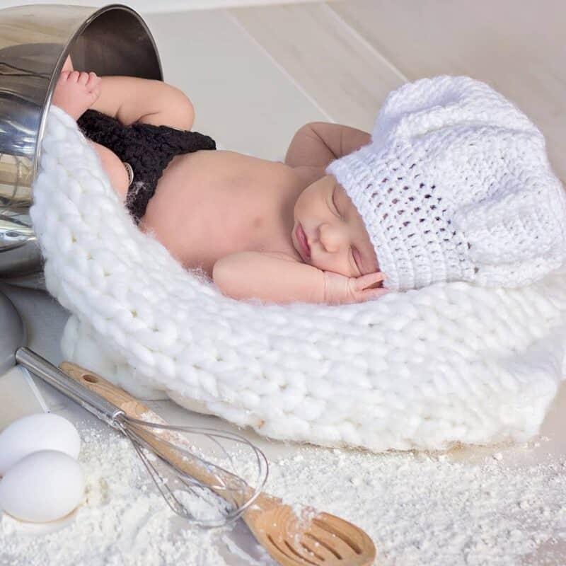 A newborn wearing a chef hat is laying in a bowl of flour.