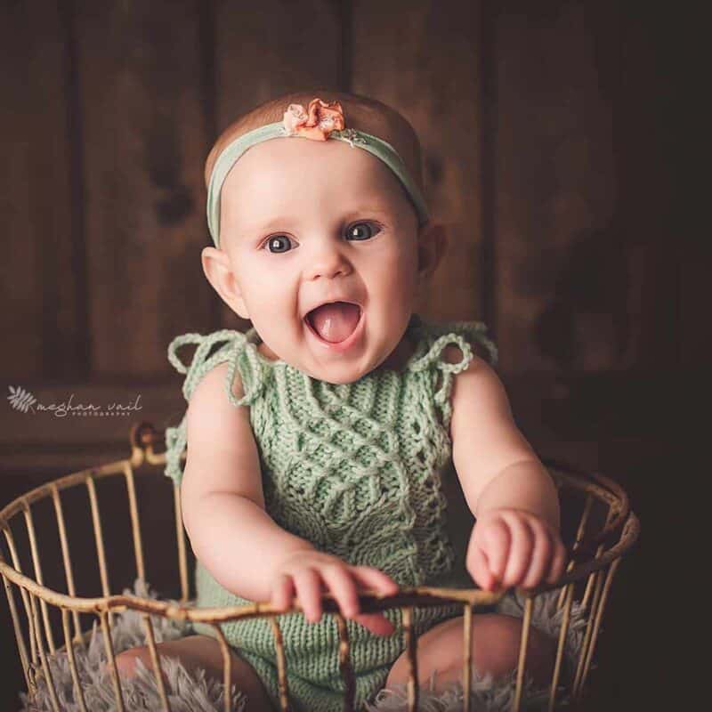 A baby wearing a Vivian Knit Romper pattern sits in a basket and laughs.