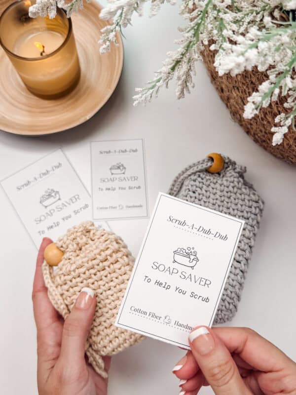 Two hands hold a beige knitted soap saver with a product tag. Nearby are a gray soap saver crochet bag, additional product tags, a lit candle, and a floral arrangement.