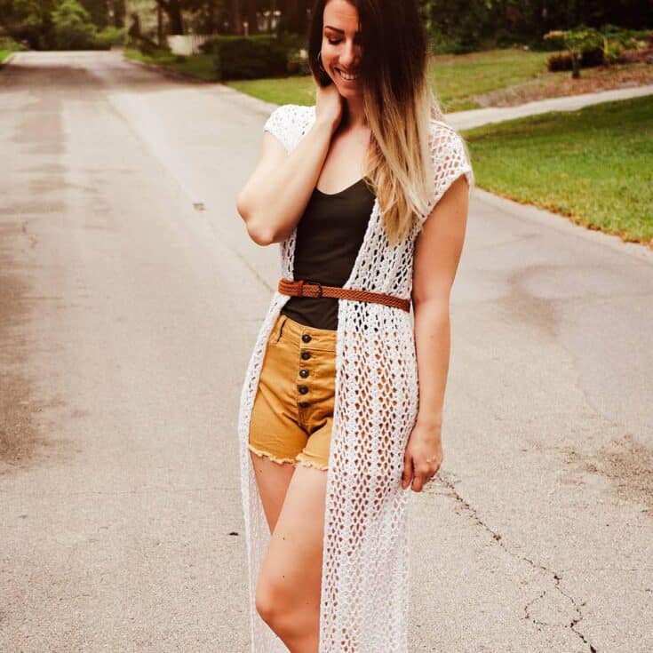 A woman wearing a long crochet duster and tan shorts.