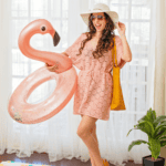 Woman wearing a pink Hexagon Cover-Up Crochet Pattern beach cover-up, sunglasses, and sunhat, holding a pink flamingo pool float and a yellow tote bag, standing on a beach towel indoors.