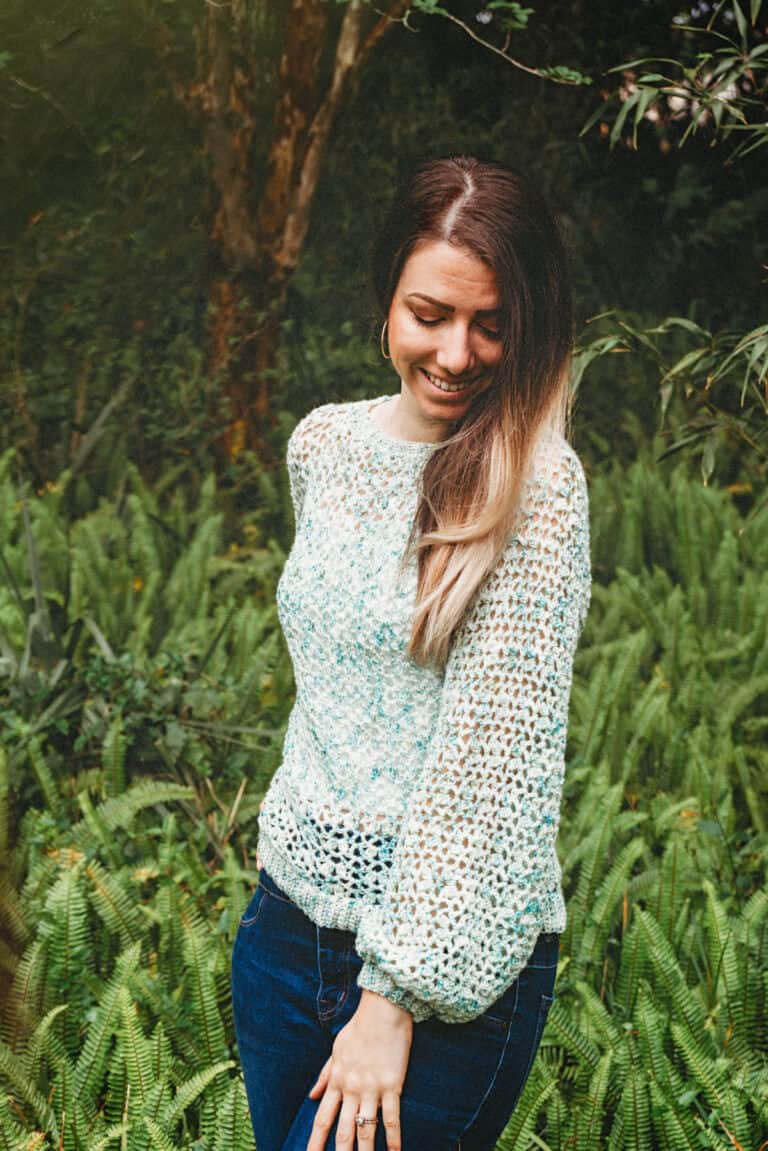 A woman smiling and looking down, wearing a crochet summer top pattern, standing against a backdrop of lush greenery.