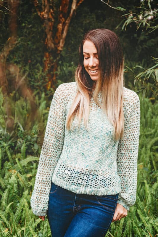 Woman smiling with closed eyes, wearing a crochet summer top, standing in front of greenery.