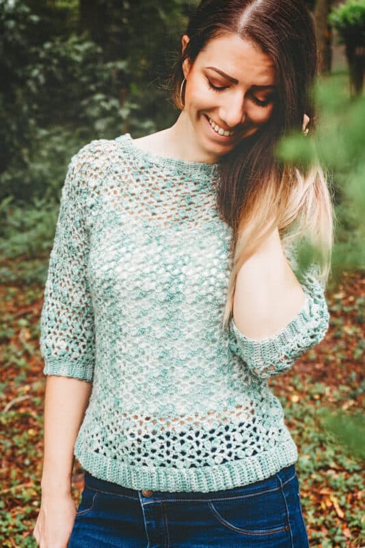 A woman smiling with closed eyes, wearing a hand-crochet summer top pattern and jeans, standing in a wooded area.