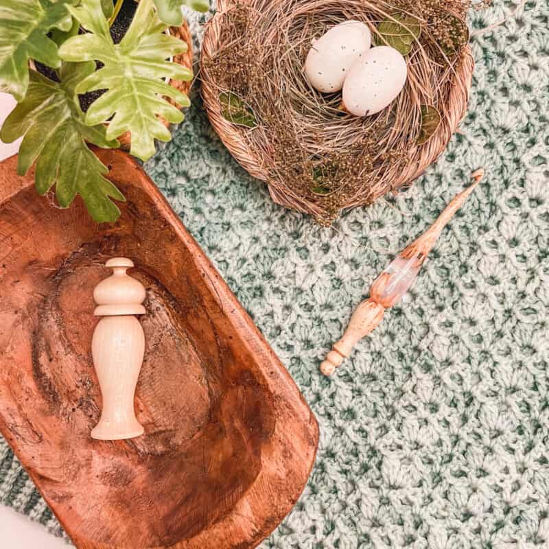 A crochet summer top pattern rests on textured fabric next to a wooden bowl and a nest with two eggs, with a green plant partially in view.