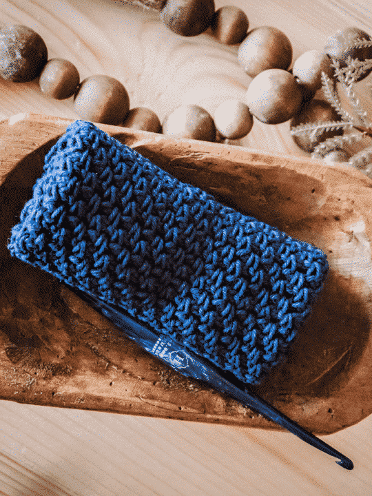 A blue crochet washcloth next to a wood bowl containing a crochet hook and decorative beads.