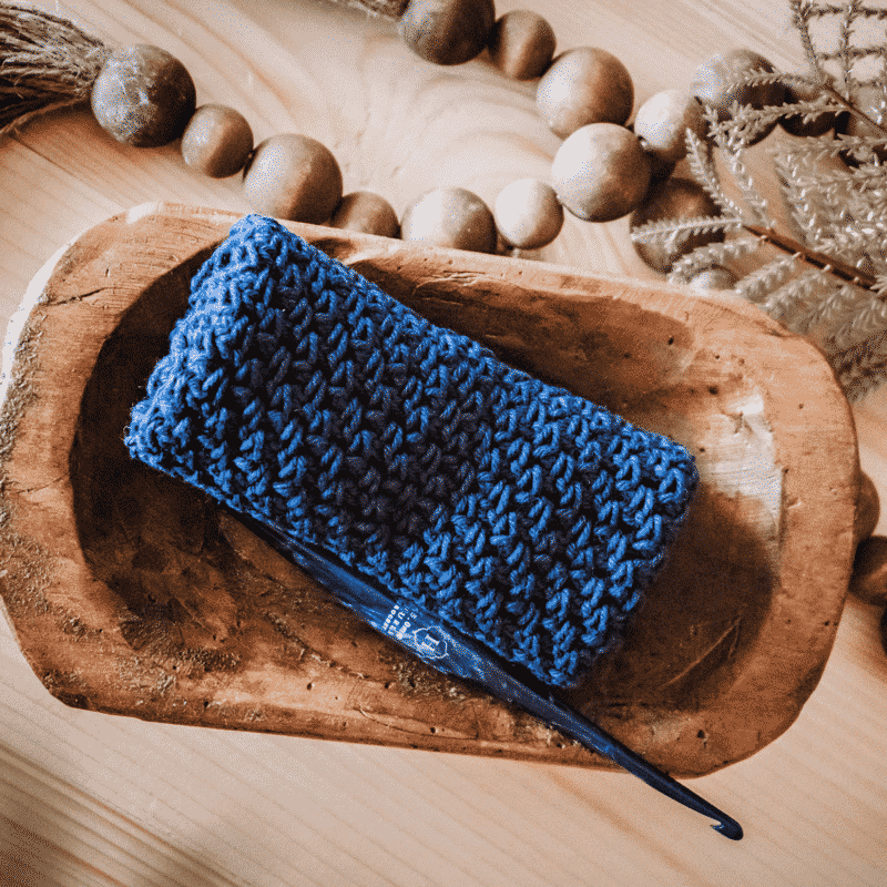 A blue crochet washcloth next to a wood bowl containing a crochet hook and decorative beads.