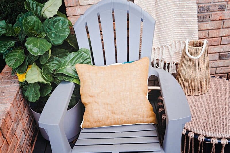 A pillow sitting on a chair next to a plant. Yarn is the pillow burlap.