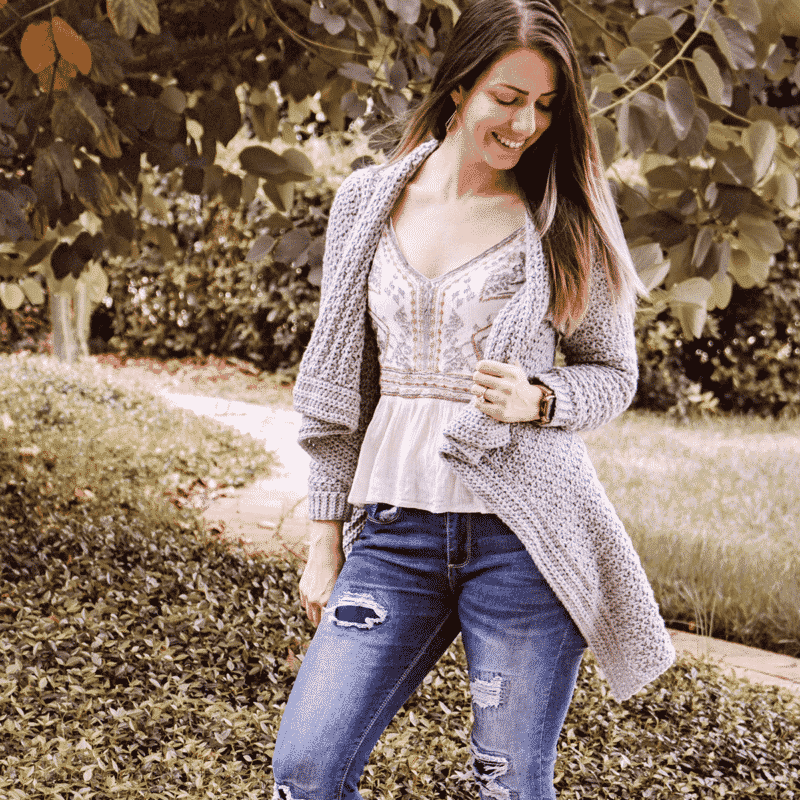 a girl standing near a tree and grass wearing a crochet grey cardigan